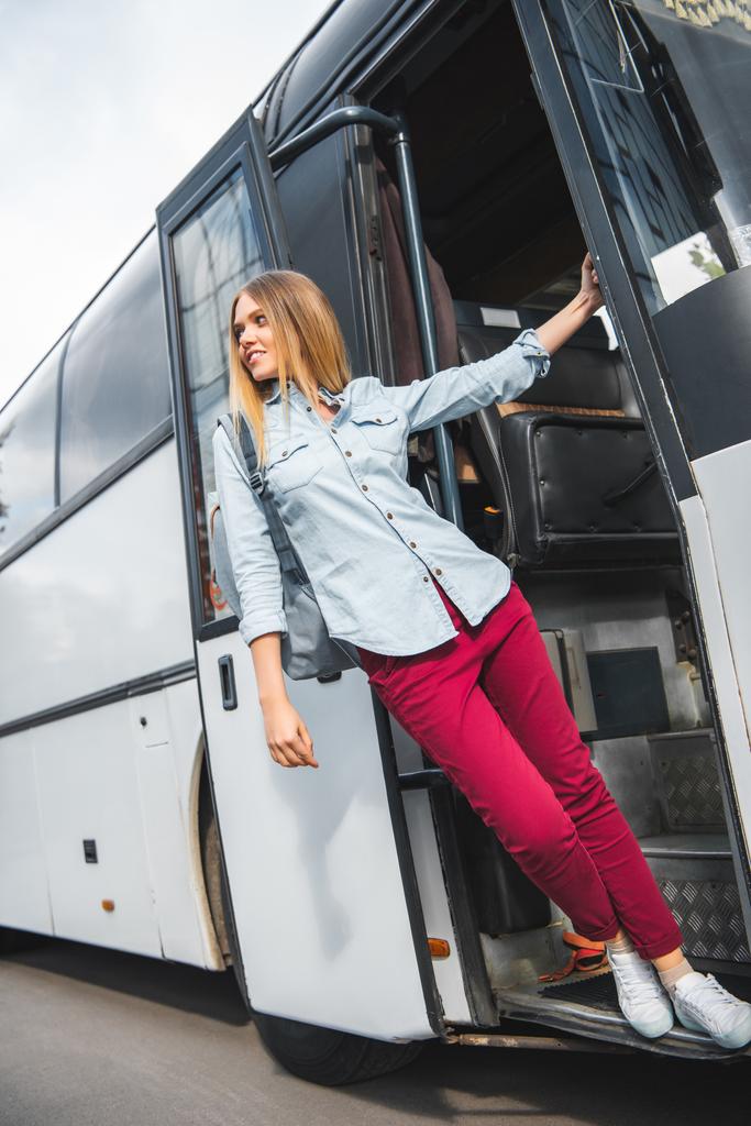 lage hoekmening van vrouwelijke toerist met rugzak poseren in de buurt van reizen bus op straat - Foto, afbeelding