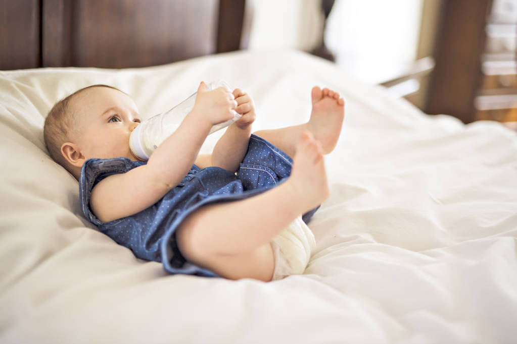Mooie babymeisje drinkt water uit een fles liggend op bed - Foto, afbeelding