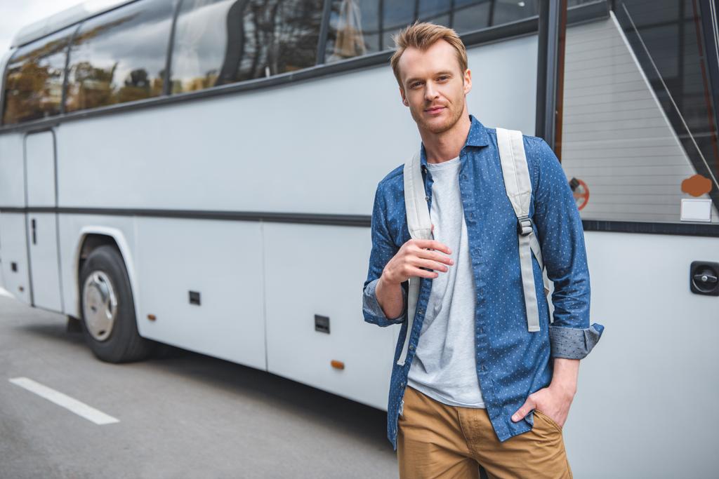 volwassen man met rugzak poseren in de buurt van reizen bus op straat - Foto, afbeelding