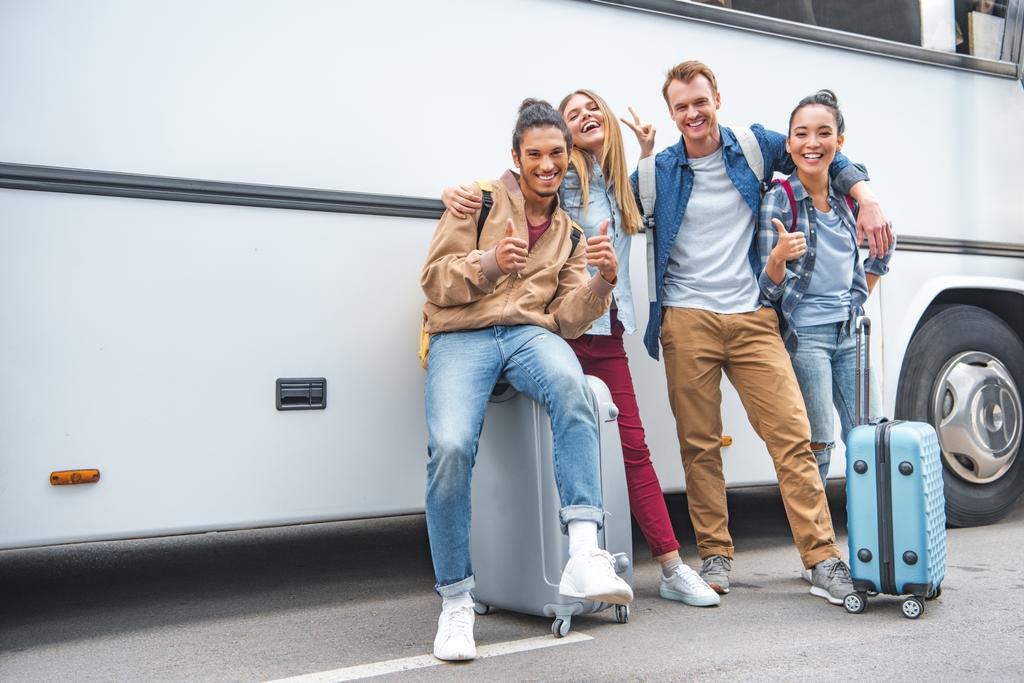 πολυεθνική τους φίλους χαμογελώντας με τροχήλατες αποσκευές κάνει χειρονομίες ειρήνης κοντά ταξιδιού λεωφορείων και μπράβο - Φωτογραφία, εικόνα