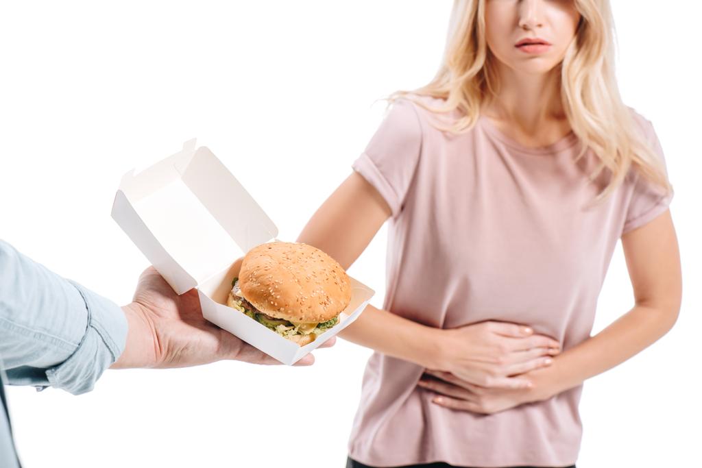 abgeschnittenes Bild der blonden Frau zeigt Bauchschmerzen, Mann schlägt ungesunden Burger isoliert auf weiß vor - Foto, Bild
