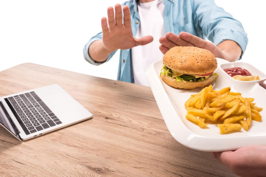 bijgesneden afbeelding van man verwerpen ongezonde hamburger en friet op tafel geïsoleerd op wit - Foto, afbeelding