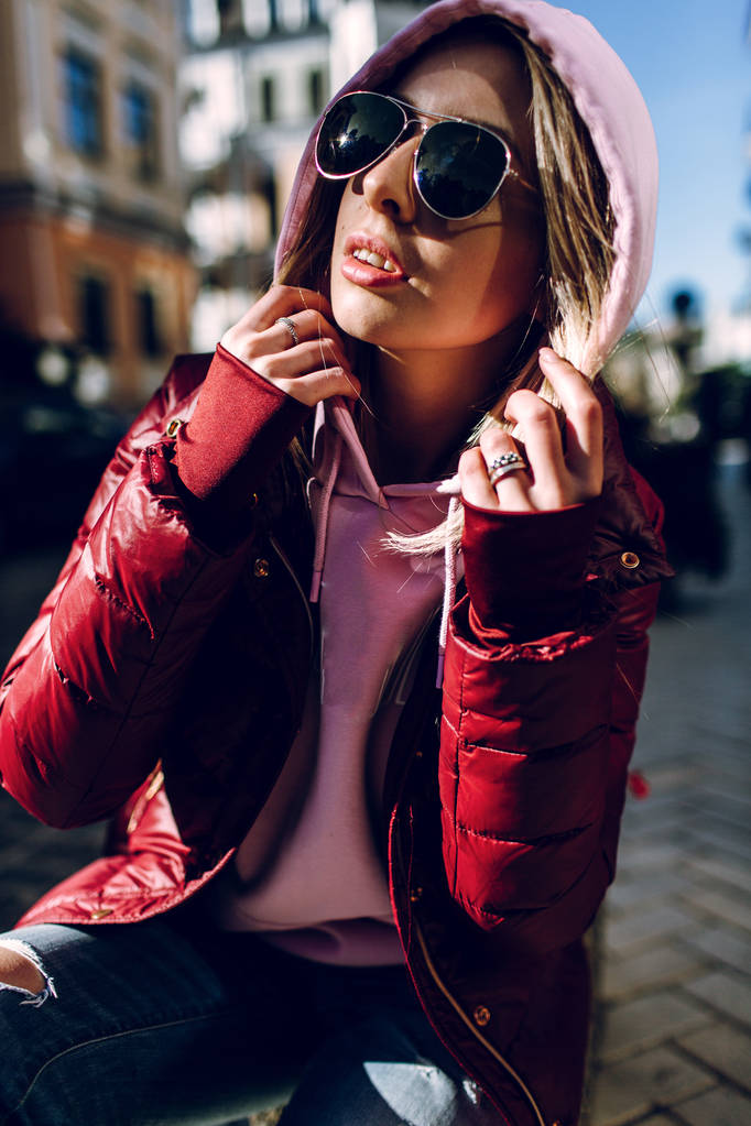 Concepto de moda callejera: retrato de una hermosa chica sentada afuera, vaqueros azules, gafas de sol, luces duras, chaqueta roja, clima otoñal.
 - Foto, Imagen