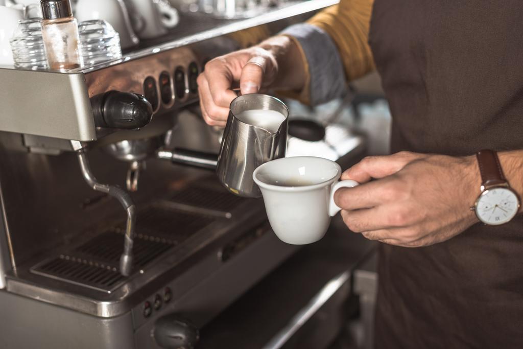 bijgesneden schot van barista in schort gieten melk in koffie bij de voorbereiding van het in café - Foto, afbeelding