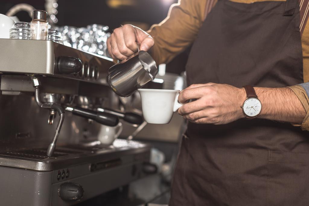 cropped shot of barista in apron pouring milk into coffee while preparing it in restaurant - Photo, Image