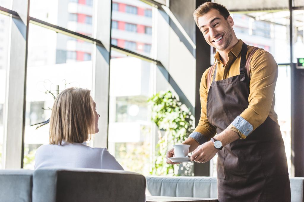wide smiling young waiter serving cup of coffee for female client at cafe - Photo, Image
