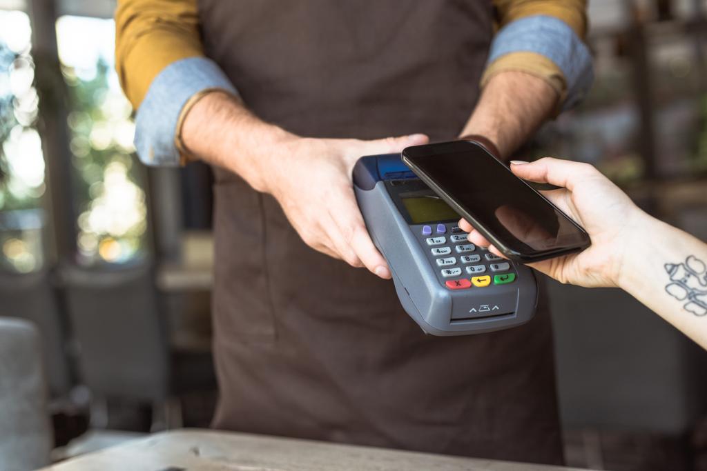 Oříznout záběr číšník držení platební terminál zároveň zákazník dělá bezkontaktní nákupu smartphonu v kavárně - Fotografie, Obrázek