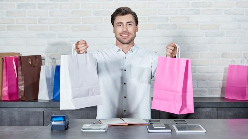 красивый молодой продавец, держащий сумки с покупками и улыбающийся перед камерой в магазине
  - Фото, изображение