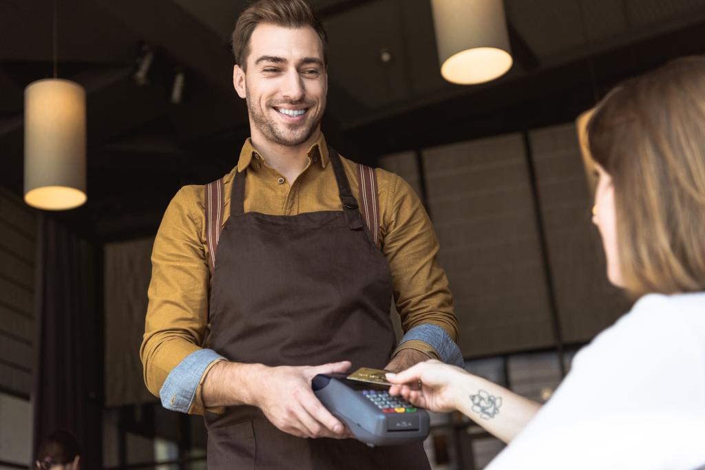 giovane cameriere sorridente che tiene terminale di pagamento mentre il cliente fa acquisto senza contatto con carta di credito in caffè
 - Foto, immagini