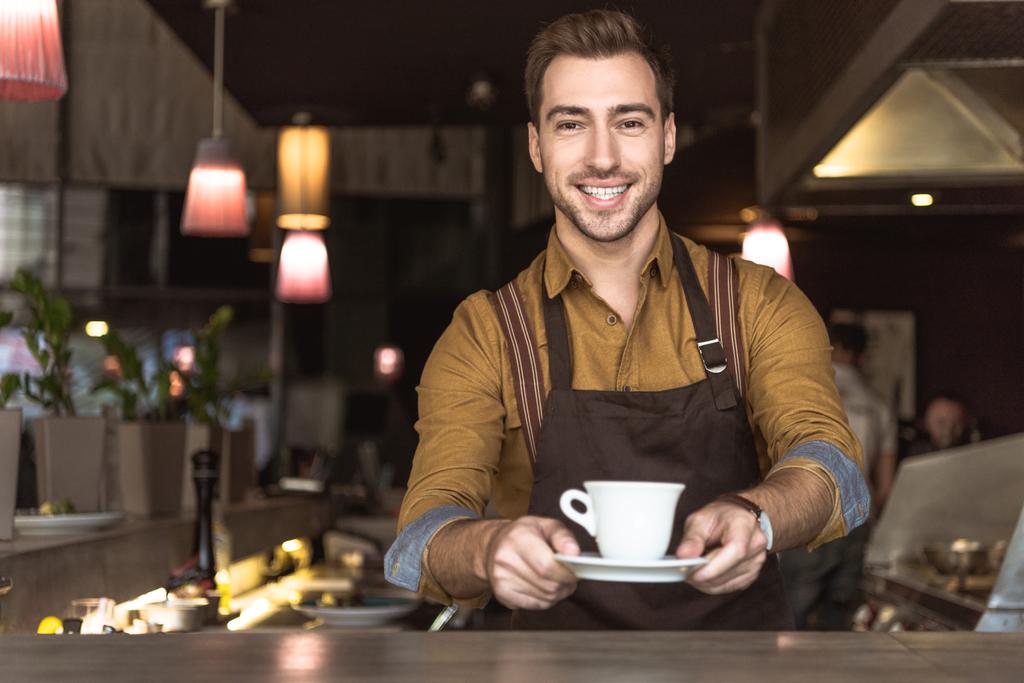 mosolygó fiatal barista gazdaság csésze kávét, és keresi a kamera - Fotó, kép