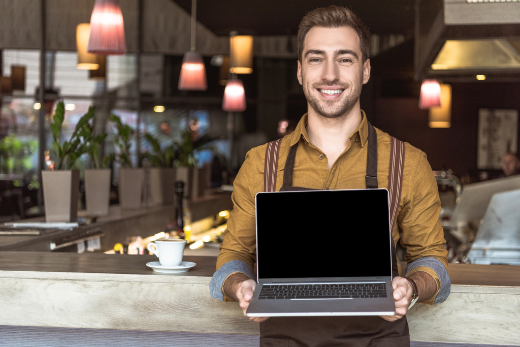счастливый молодой официант держит ноутбук с пустым экраном в кафе
 - Фото, изображение