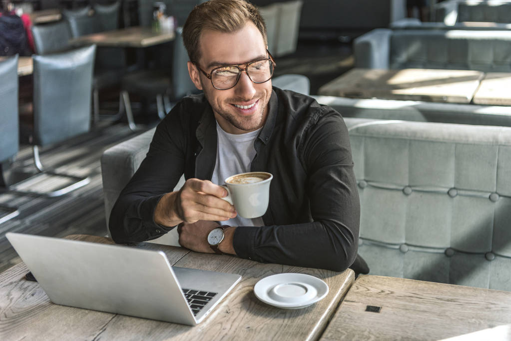 красивый молодой фрилансер с чашкой кофе и ноутбуком, сидящий в кафе
 - Фото, изображение