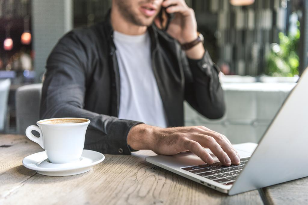 обрезанный снимок молодого фрилансера с чашкой кофе, работающего с ноутбуком в кафе и разговаривающего по телефону
 - Фото, изображение
