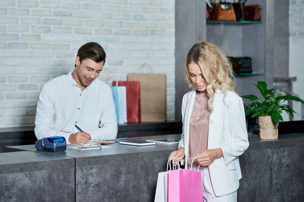 jovem mulher olhando em sacos de compras enquanto vendedor tomar notas na loja
 - Foto, Imagem