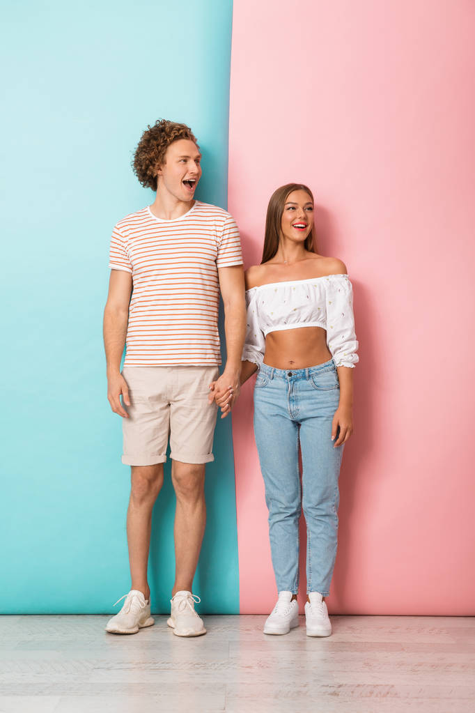 2 つの色付きの背景の上に立って、離れて見て幸せな若いカップルの完全な長さの肖像画 - 写真・画像