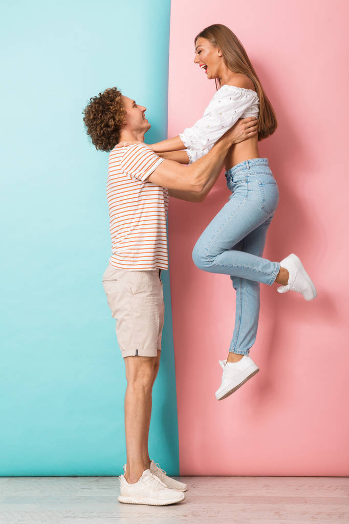 Ganzkörperporträt eines glücklichen jungen Paares, das vor zweifarbigem Hintergrund steht, einander ansieht, seine Freundin hochhebt - Foto, Bild