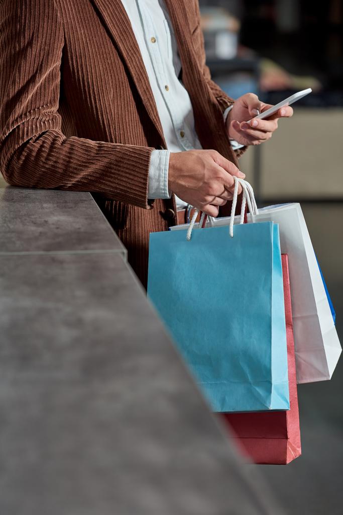 περικοπεί shot του ανθρώπου, κρατώντας τσάντες αγορών και χρησιμοποιώντας το smartphone στο κατάστημα - Φωτογραφία, εικόνα