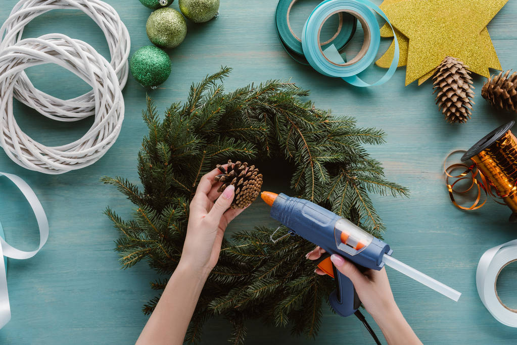 cropped shot of woman decorating handmade christmas wreath with pine cone on blue wooden surface - Photo, Image