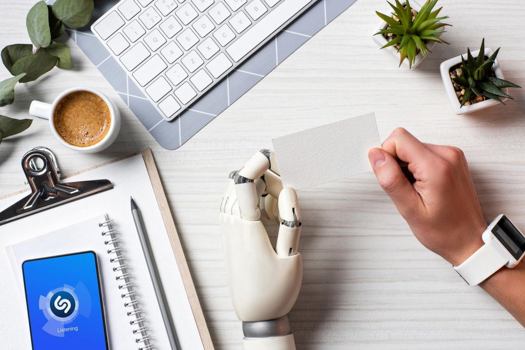 Ausgeschnittenes Bild eines Geschäftsmannes mit Cyborg-Hand und Smartwatch mit leerer Visitenkarte am Tisch mit Smartphone mit Shazam auf dem Bildschirm im Büro  - Foto, Bild