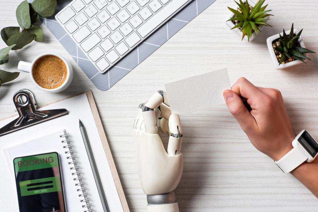 Ausgeschnittenes Bild eines Geschäftsmannes mit Cyborg-Hand und Smartwatch mit leerer Visitenkarte am Tisch mit Smartphone mit Buchung auf dem Bildschirm im Büro  - Foto, Bild
