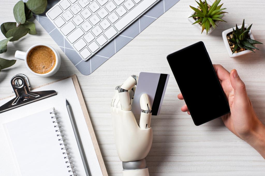 обрезанный образ бизнесмена с протезной рукой, держащего кредитную карту и использующего смартфон с чистым экраном за столом в офисе
  - Фото, изображение