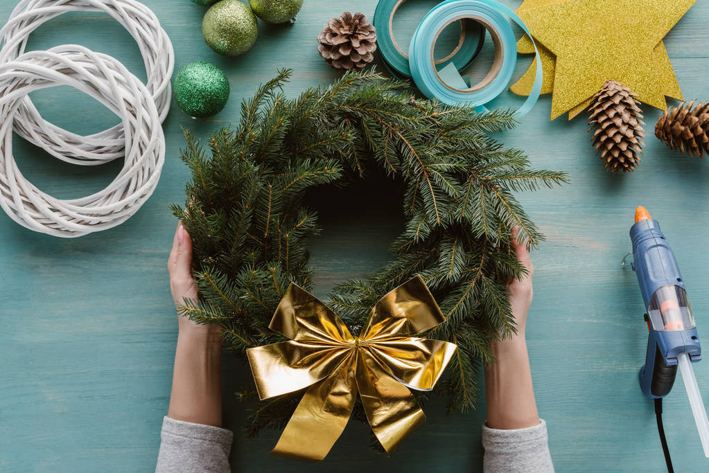 cropped shot of woman holding handmade pine tree wreath with golden bow and decorations on blue wooden tabletop - Photo, Image