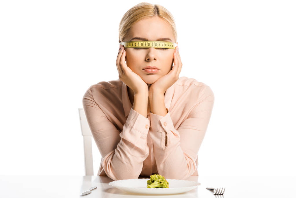 aantrekkelijke vrouw met meetlint op ogen zitten in de buurt van stukje broccoli op plaat geïsoleerd op wit - Foto, afbeelding