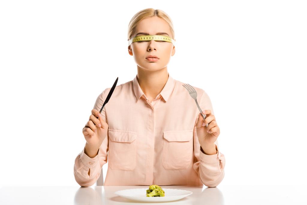 aantrekkelijke vrouw met meetlint op ogen houden vork en mes, stukje broccoli op plaat geïsoleerd op wit - Foto, afbeelding