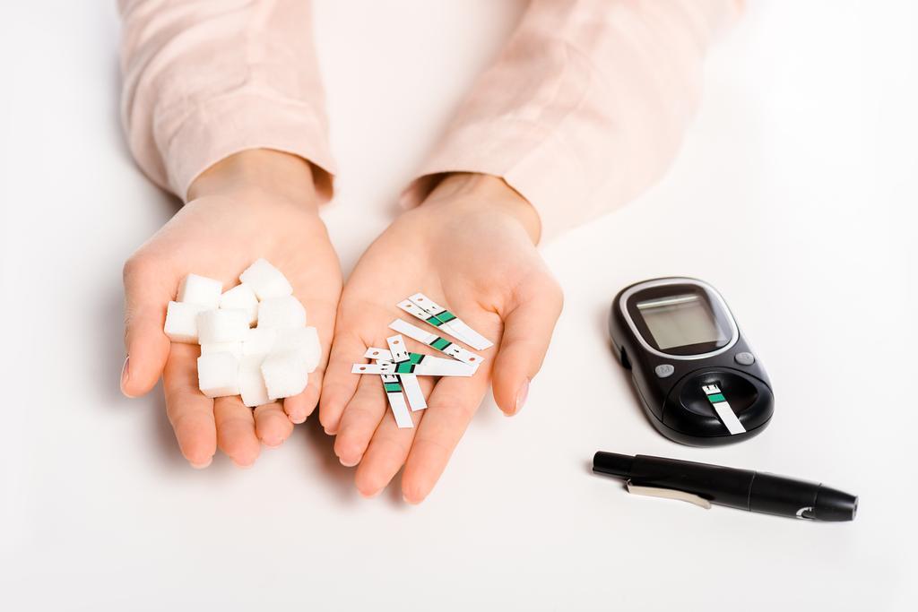 περικομμένη εικόνα γυναίκας που κρατά glucometer λωρίδες και ραφιναρισμένη ζάχαρη που απομονώνονται σε λευκό, έννοια του διαβήτη - Φωτογραφία, εικόνα