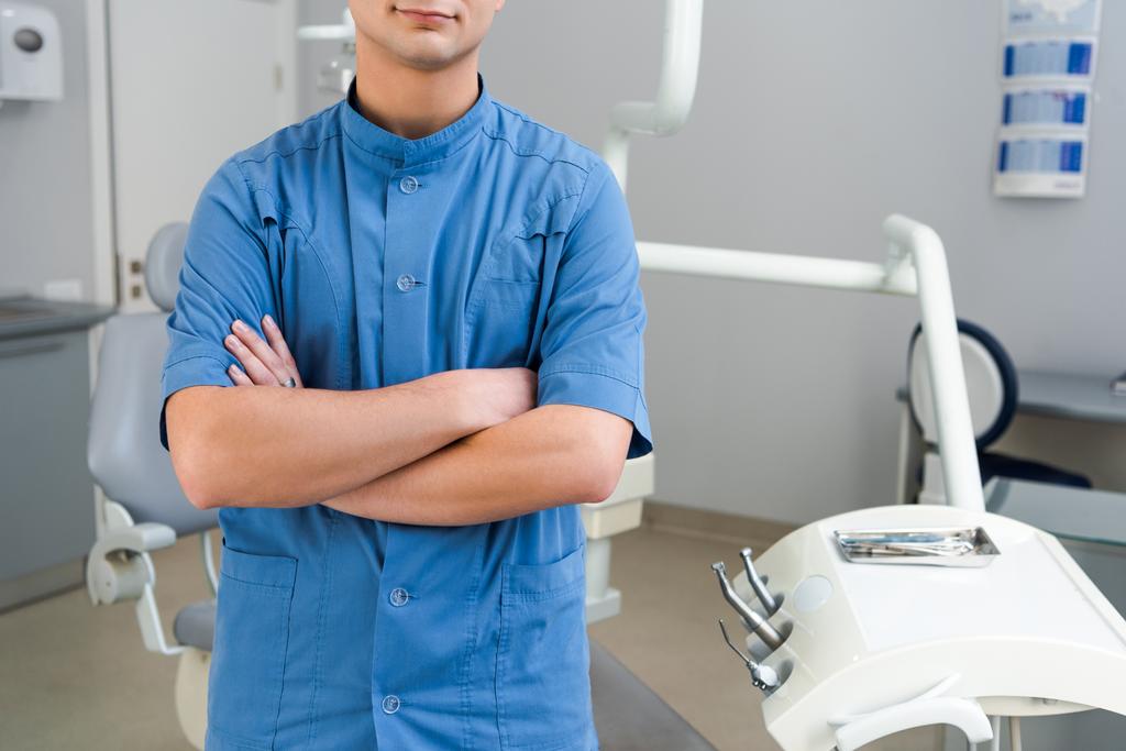 schot van jonge tandarts bijgesneden in blauwe uniform met gekruiste armen in kantoor - Foto, afbeelding