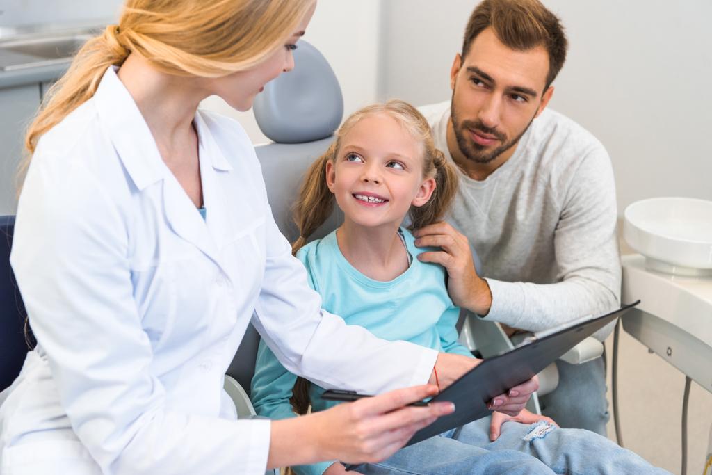 jovem dentista mostrando prancheta com diagnóstico para pai e filha
 - Foto, Imagem