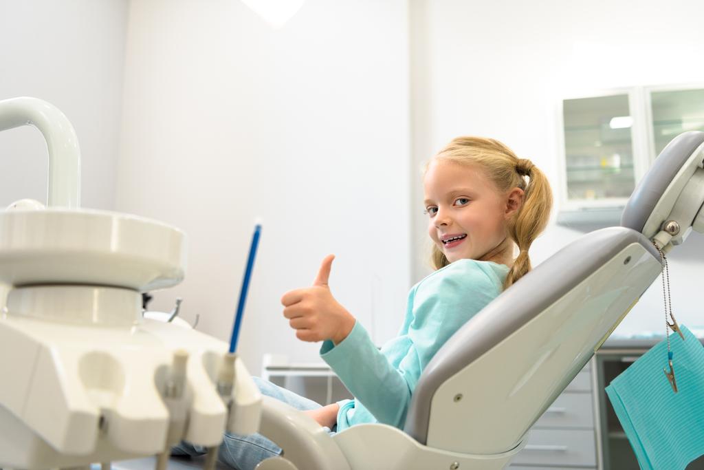 Niño feliz sentado en la silla dental en el consultorio del dentista y mostrando el pulgar hacia arriba - Foto, imagen