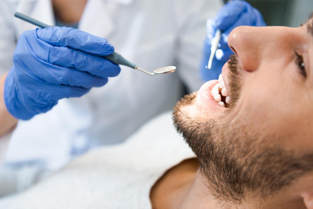 обрезанный снимок стоматолога, осматривающего зубы красивого улыбающегося клиента
 - Фото, изображение