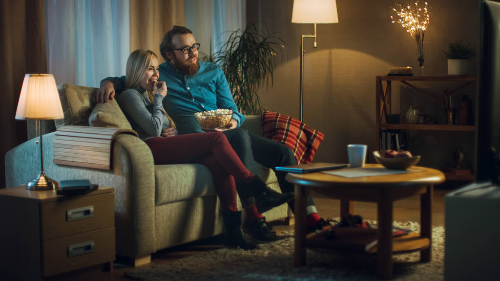 Az este férfi és a nő ül a kanapén tévénézés és étkezési Popcorn. Nappali egy hangulatos. - Fotó, kép