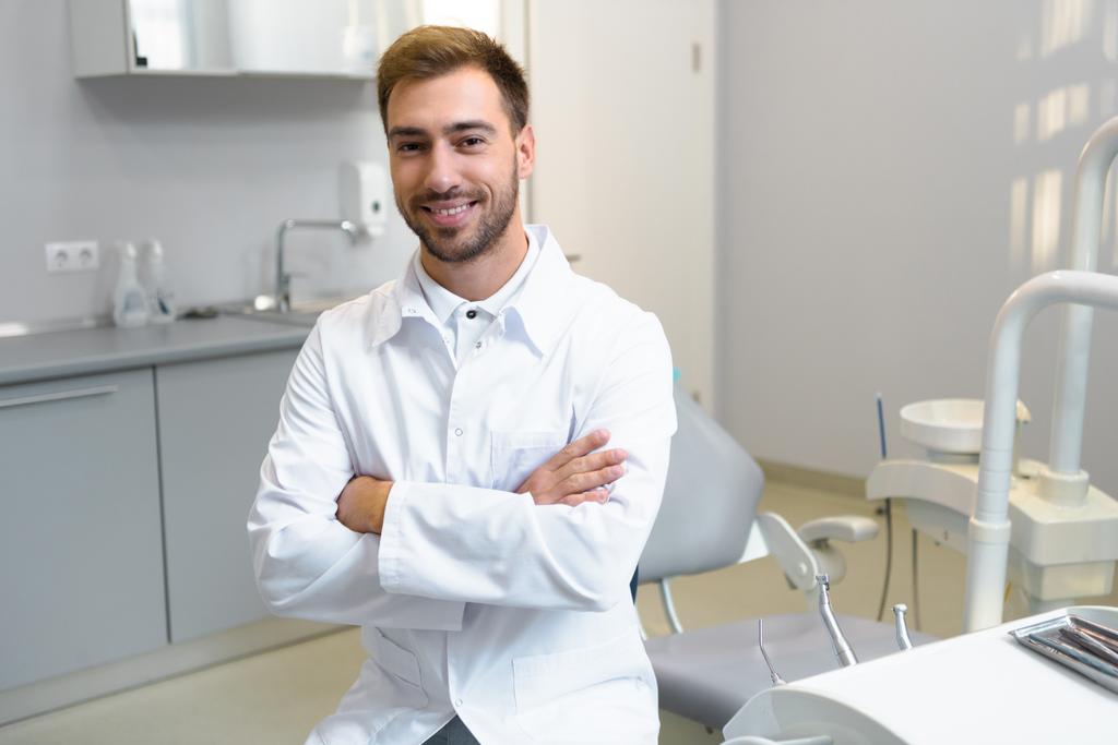 šťastný mladý zubař s překřížením rukou při pohledu na fotoaparát v úřadu - Fotografie, Obrázek