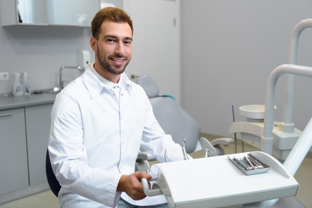 sorridente jovem dentista de casaco branco olhando para a câmera no escritório
 - Foto, Imagem