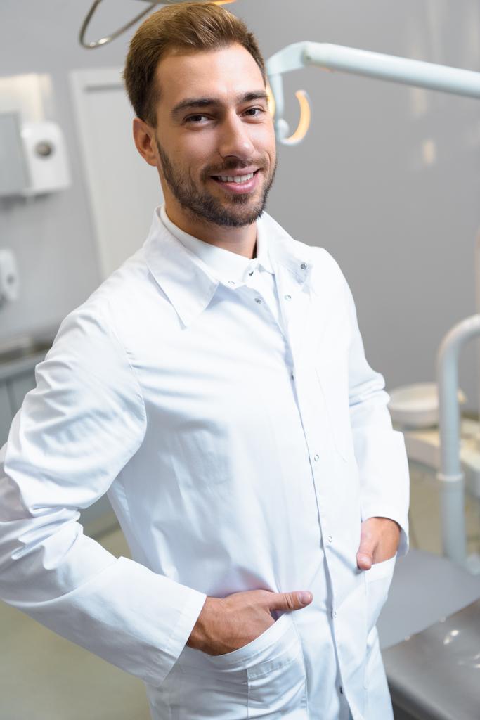 красивий молодий стоматолог у білому пальто дивиться на камеру в офісі
 - Фото, зображення