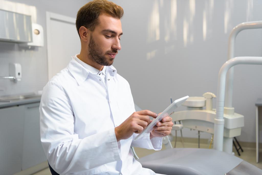 knappe jonge tandarts in vacht tablet gebruiken in office - Foto, afbeelding