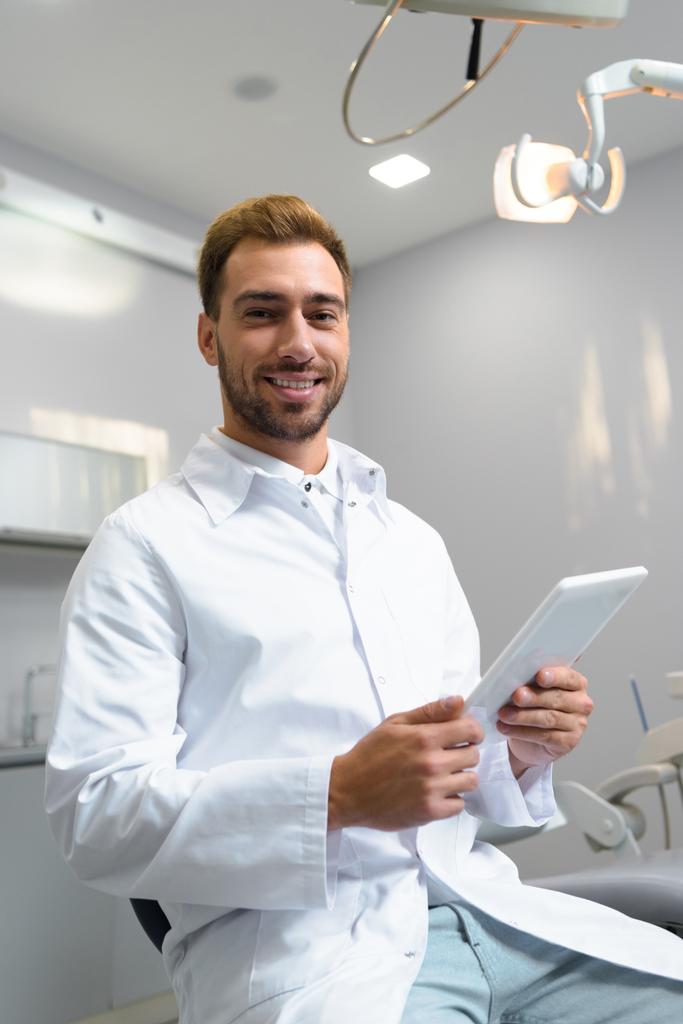 šťastný mladý zubař v kabátě pomocí tabletu v kanceláři - Fotografie, Obrázek