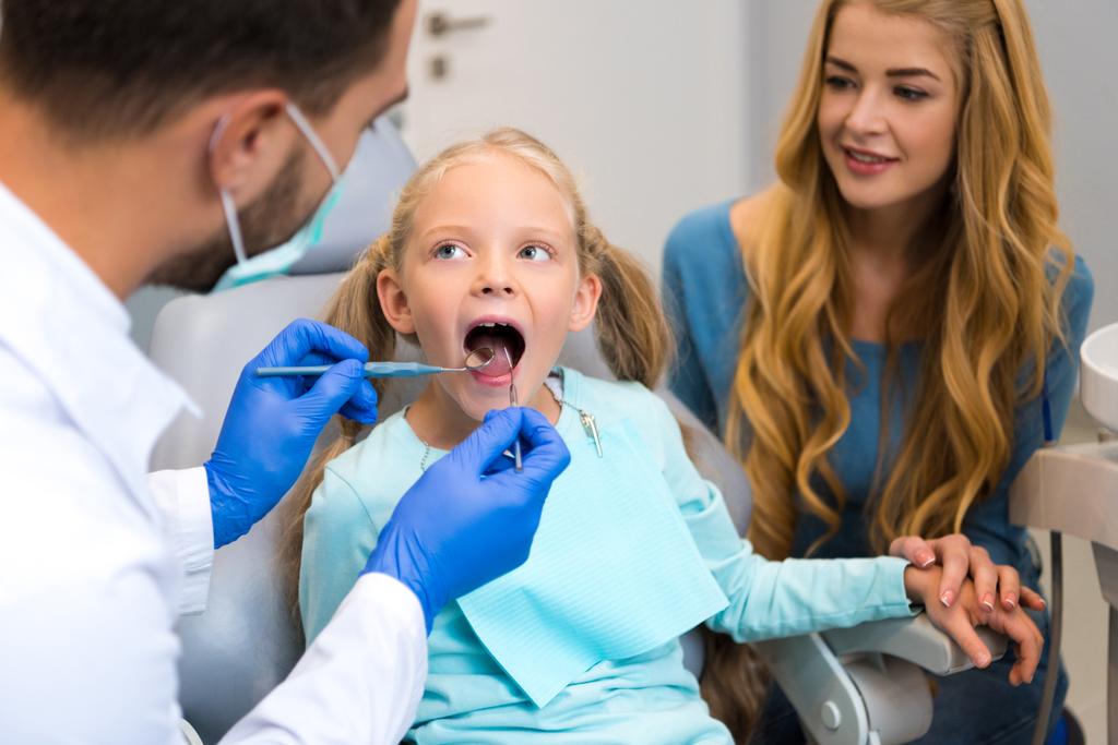dentista examinando los dientes de un niño pequeño mientras la madre está sentada cerca de ella
 - Foto, Imagen