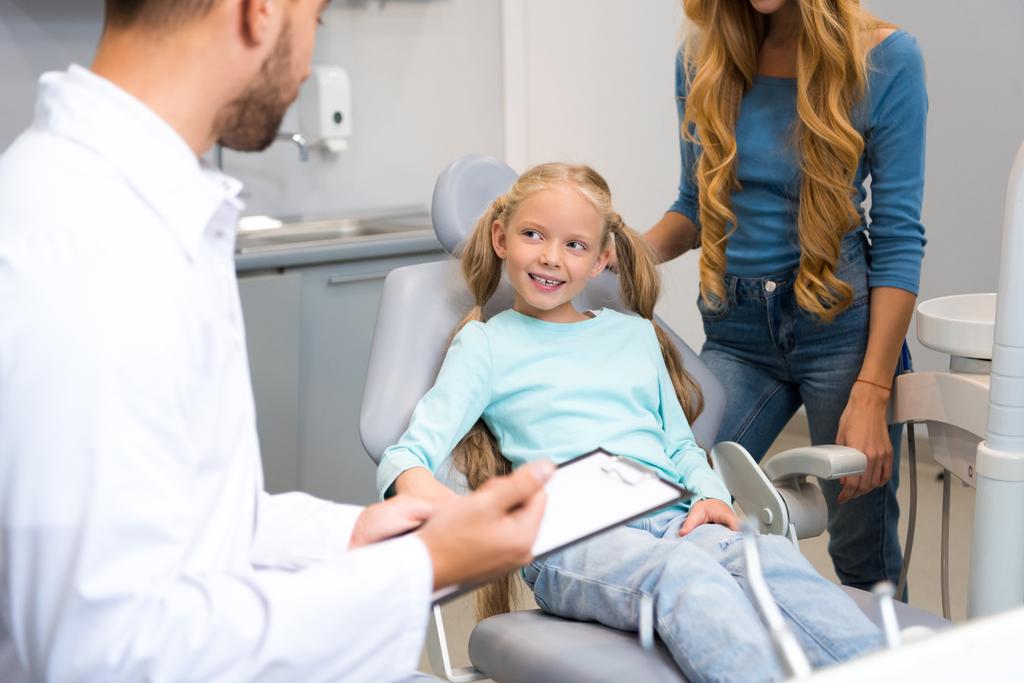 Zahnarzt mit Klemmbrett spricht mit kleinem Kind, während die Mutter neben ihr zur Unterstützung steht - Foto, Bild