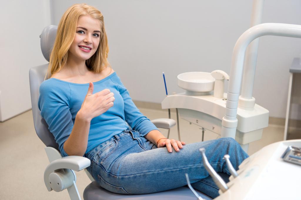 šťastná mladá žena sedí v Zubařské křeslo v ordinaci a ukazuje palec - Fotografie, Obrázek