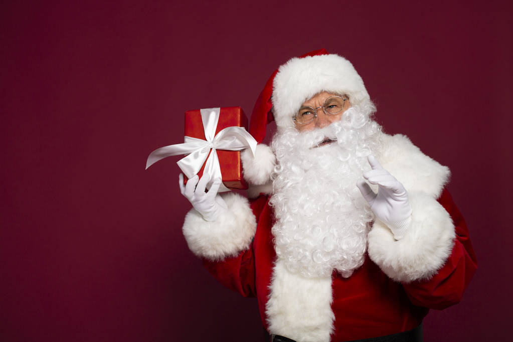 πορτρέτο του ανθρώπου στη Santa ρήτρα κοστούμι κρατώντας κουτί δώρου χαρτί και βλέπουν τα φωτογραφικών μηχανών σε κόκκινο φόντο, Χριστούγεννα και νέο έτος έννοια  - Φωτογραφία, εικόνα