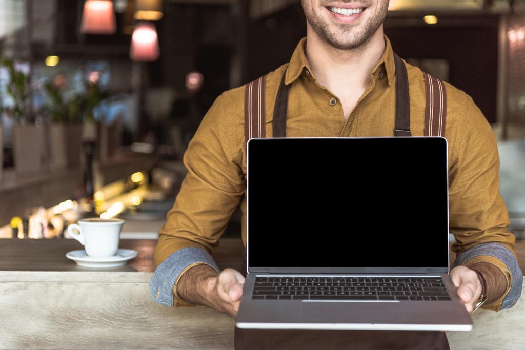 カフェで空白の画面を持つノート パソコンを持って笑顔の若いウェイターのショットをトリミング - 写真・画像