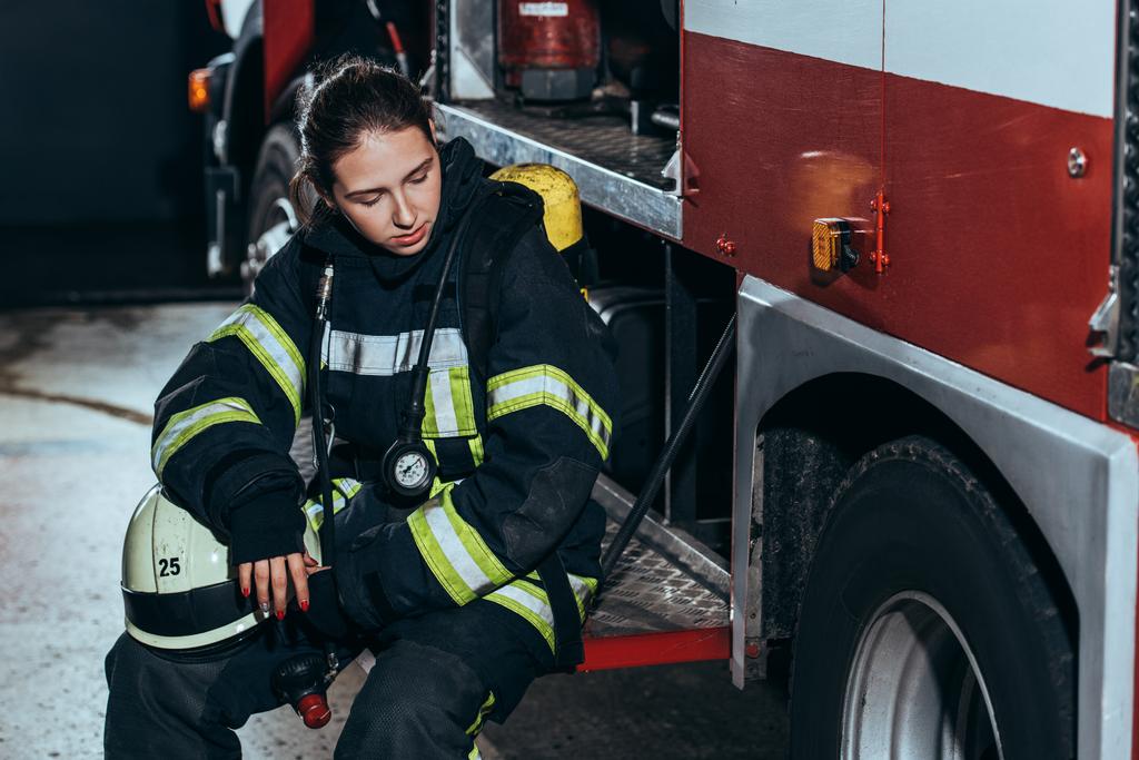 mujer bombero cansado en uniforme con casco sentado en el camión en la estación de bomberos
 - Foto, Imagen