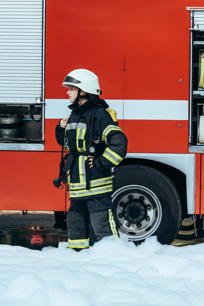 Feuerwehrfrau in Schutzuniform steht in Schaumstoff auf der Straße, dahinter rotes Feuerwehrauto - Foto, Bild