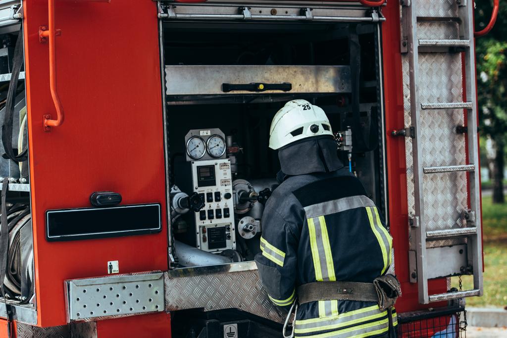 Rückansicht eines Feuerwehrmannes in Uniform und Helm, der am Feuerwehrauto auf der Straße steht - Foto, Bild