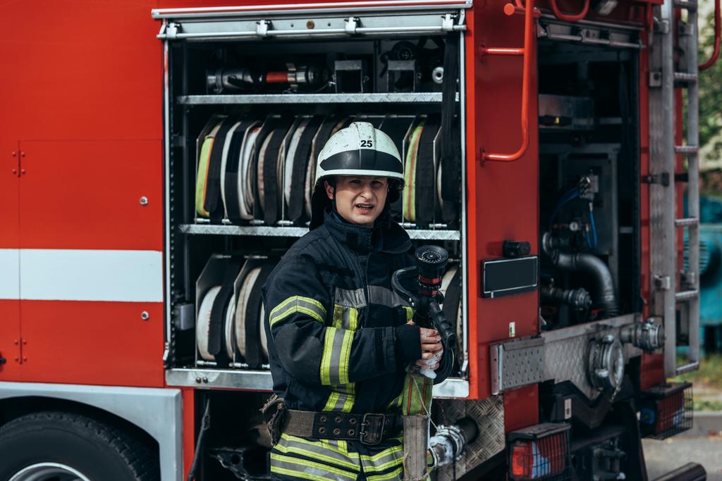 Porträt eines Feuerwehrmannes in Schutzuniform und Helm, der mit Wasserschläuchen auf der Straße am LKW steht - Foto, Bild