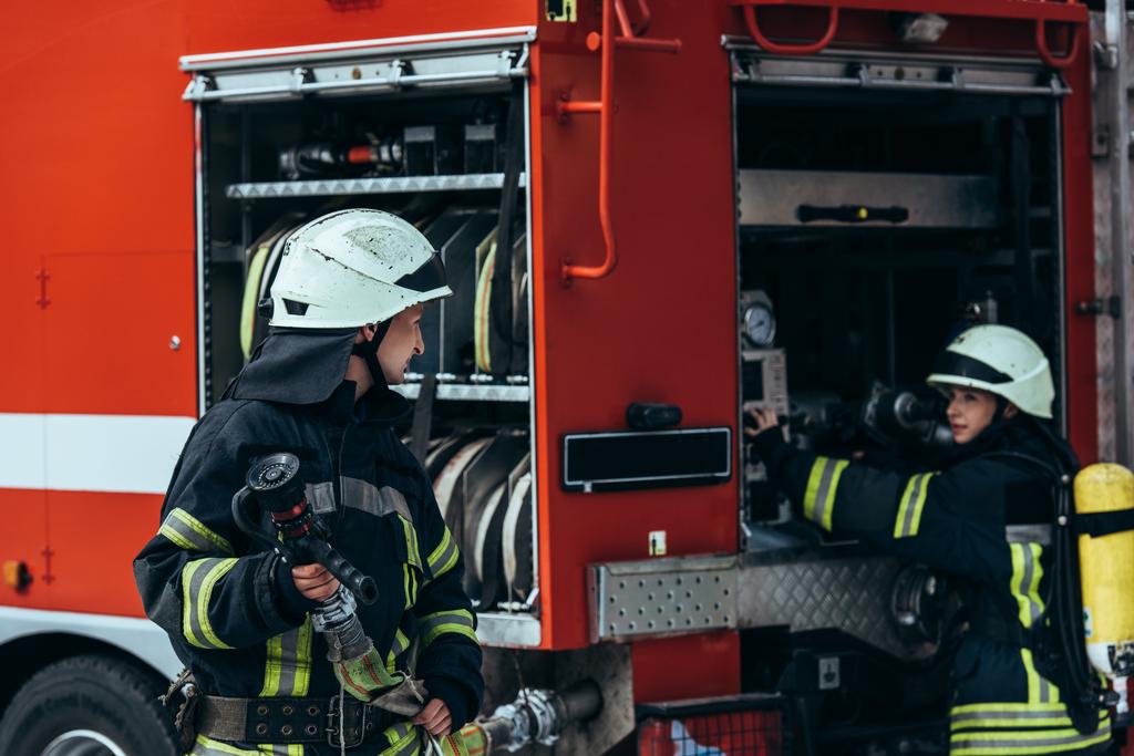 bombeiro masculino em uniforme segurando mangueira de água enquanto colega verificando equipamentos em caminhão na rua
 - Foto, Imagem