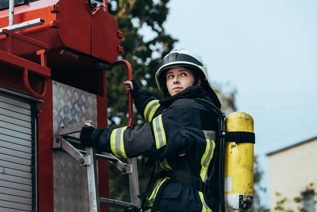 vrouwelijke brandweerman met brandblusser op terug staande op de brandweerwagen op straat - Foto, afbeelding